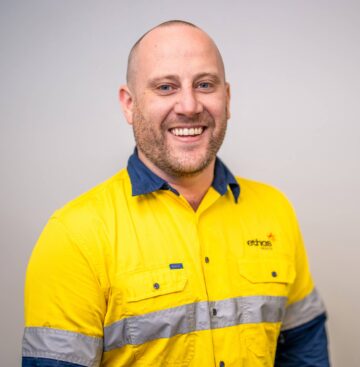 Friendly healthcare professional wearing bright yellow Ethos Health uniform; indoor setting.
