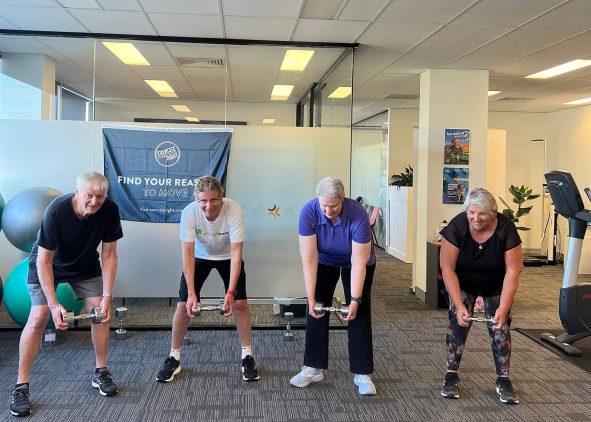 Group of four older adults participating in a strength training session at a gym, lifting dumbbells and barbells while smiling.