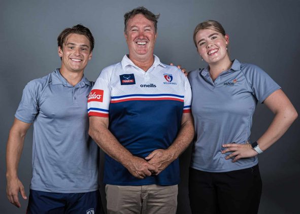 Three smiling individuals posing against a grey background, with the central figure wearing a Newcastle Wildfires rugby jersey and the two individuals on either side wearing Ethos Health polo shirts.