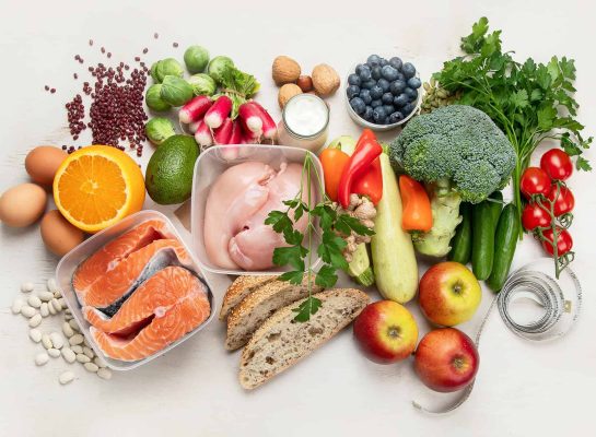Assortment of healthy foods including fresh salmon, chicken, eggs, whole grain bread, fruits, vegetables, legumes, nuts, and yogurt, arranged on a white background.