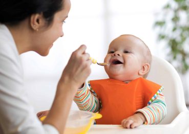 Smiling mother feeding her baby with a spoon while the baby, wearing an orange bib and a striped long-sleeve shirt, sits in a high chair.
