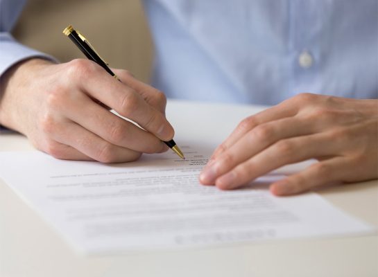 man's hands signing a document with a black and gold pen. He is wearing a light blue dress shirt, and the contract or agreement is placed on a white desk.