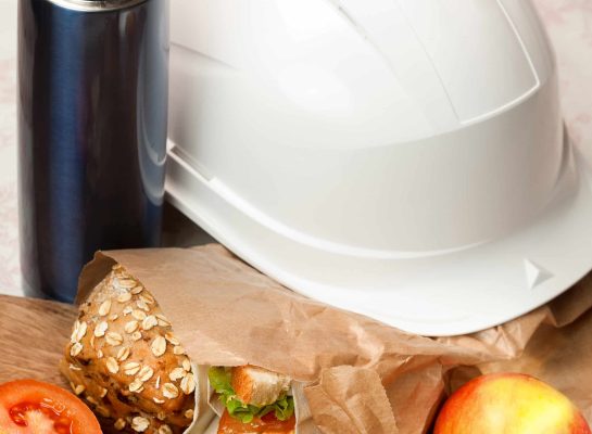 Close-up of a healthy packed lunch with a whole grain sandwich in a brown paper bag, a fresh apple, and a tomato slice next to a stainless steel thermos and a white hard hat.