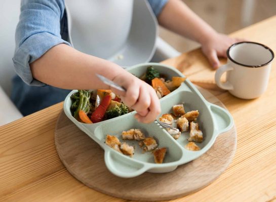 young child eating a meal from a divided plate with chicken nuggets and roasted vegetables.