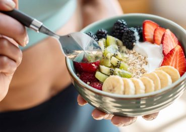 Close-up of a fit woman holding a green bowl filled with a nutritious breakfast of oats, yogurt, and fresh fruit, including bananas, strawberries, kiwi, blackberries, and raspberries.