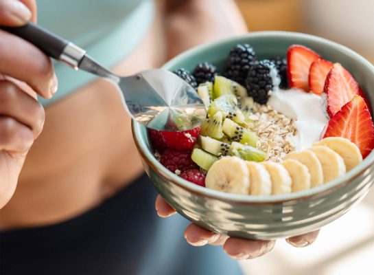 Close-up of a fit woman holding a green bowl filled with a nutritious breakfast of oats, yogurt, and fresh fruit, including bananas, strawberries, kiwi, blackberries, and raspberries.