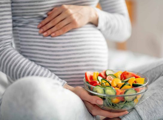 Pregnant woman in a grey striped shirt holding a glass bowl of fresh vegetable salad with cucumbers, tomatoes, and bell peppers.