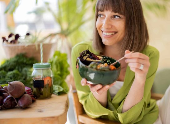 Smiling woman in a green blouse enjoying a healthy meal from a green bowl, surrounded by fresh vegetables, herbs, and a jar of infused water.