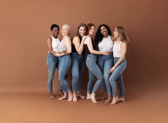 Group of six diverse women of different ages and ethnicities posing together in jeans and neutral-toned tops against a brown background.