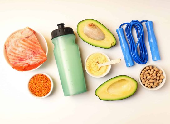 Flat lay of fitness and nutrition essentials, including fresh salmon, avocado halves, red lentils, chickpeas, protein powder, a green water bottle, and a blue jump rope