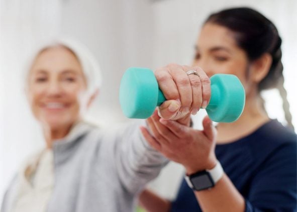 Close-up of an older woman performing a strength exercise with a light green dumbbell, assisted by a female trainer in a navy top.