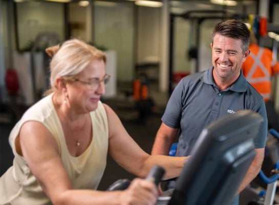 Smiling male fitness trainer in an Ethos Health uniform assisting an older woman using a stationary exercise bike in a gym.