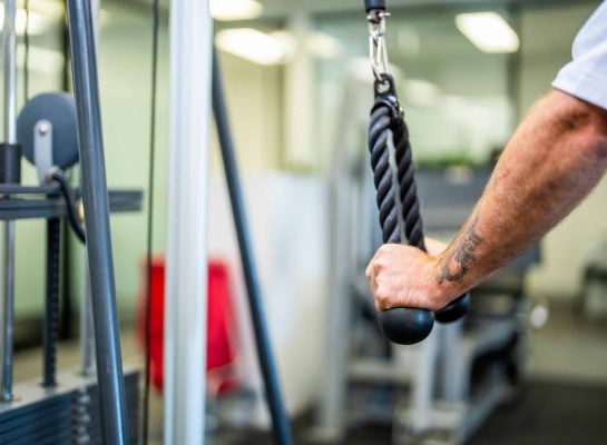 Close-up of a man's arm with a tattoo performing a tricep rope pushdown exercise on a cable machine in a gym.