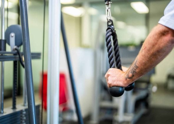 Close-up of a man's arm with a tattoo performing a tricep rope pushdown exercise on a cable machine in a gym.