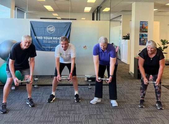 A group of four older adults participating in a strength training session at Ethos Health, holding dumbbells and barbells while bending forward.