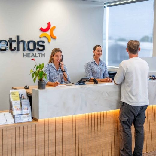Reception area of Ethos Health with two friendly receptionists in striped uniforms assisting a male visitor.